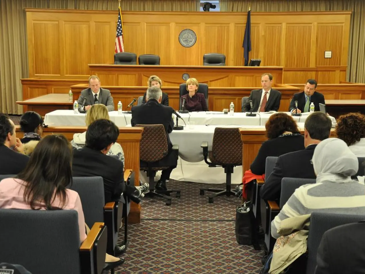This is a meeting hall where we can see a group of people sitting on chairs and also two flags of...