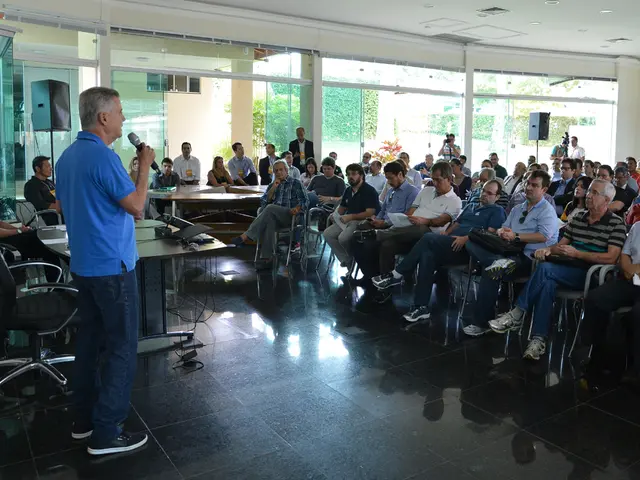 A picture of a room. This man is standing and holds a mic. On this table there is a projector and...